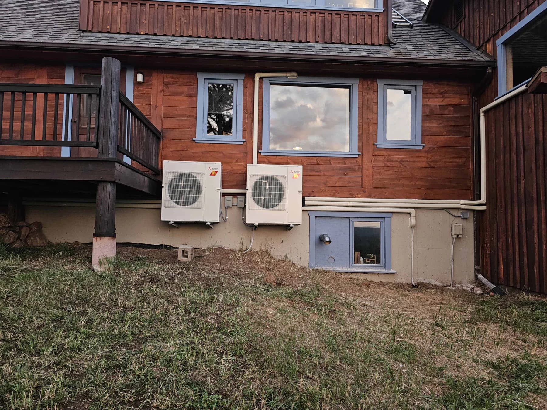 Two outdoor units beside a wood-clad house, deck and basement window